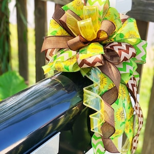 May include: A decorative bow featuring a sunflower theme, attached to a black mailbox. The bow incorporates various ribbons in shades of yellow, green, and brown. Some ribbons have sunflower patterns, while others have chevron or solid designs. The bow is attached to a white post.