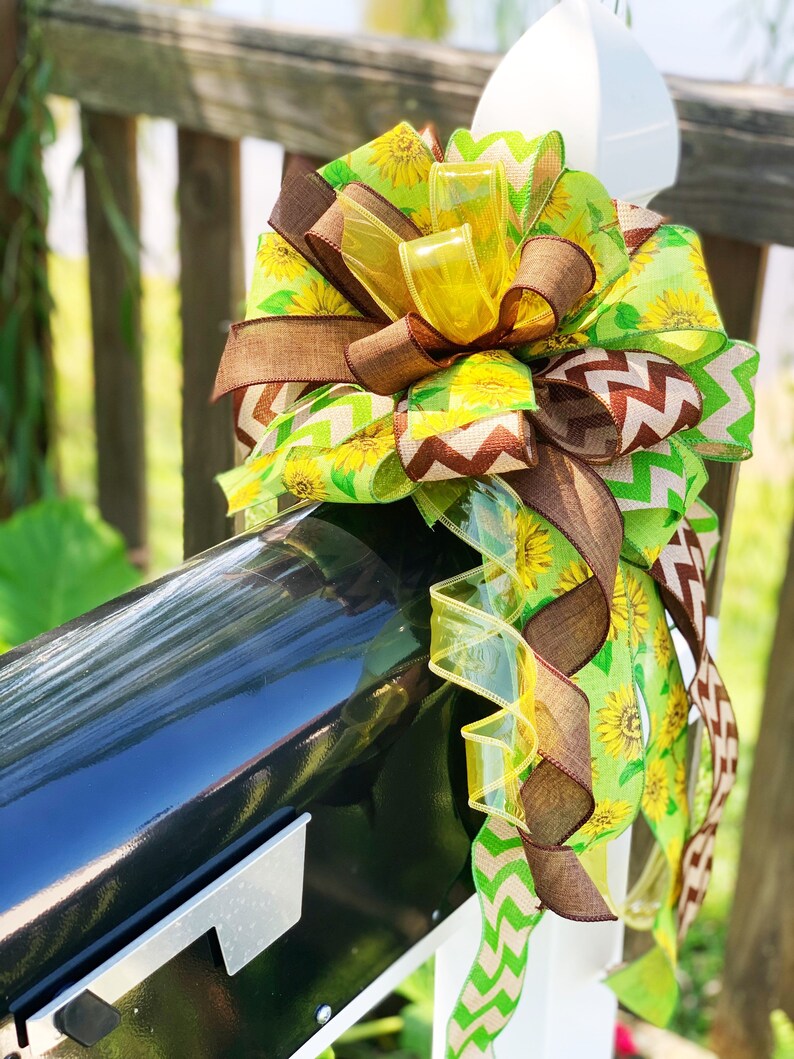 May include: A decorative bow featuring a sunflower theme, attached to a black mailbox. The bow incorporates various ribbons in shades of yellow, green, and brown. Some ribbons have sunflower patterns, while others have chevron or solid designs. The bow is attached to a white post.