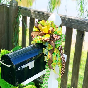 May include: A black mailbox with a decorative bow. The bow features yellow sunflowers, brown ribbons, and green chevron and solid ribbons. The mailbox is mounted on a white post, with a wooden fence and greenery in the background.