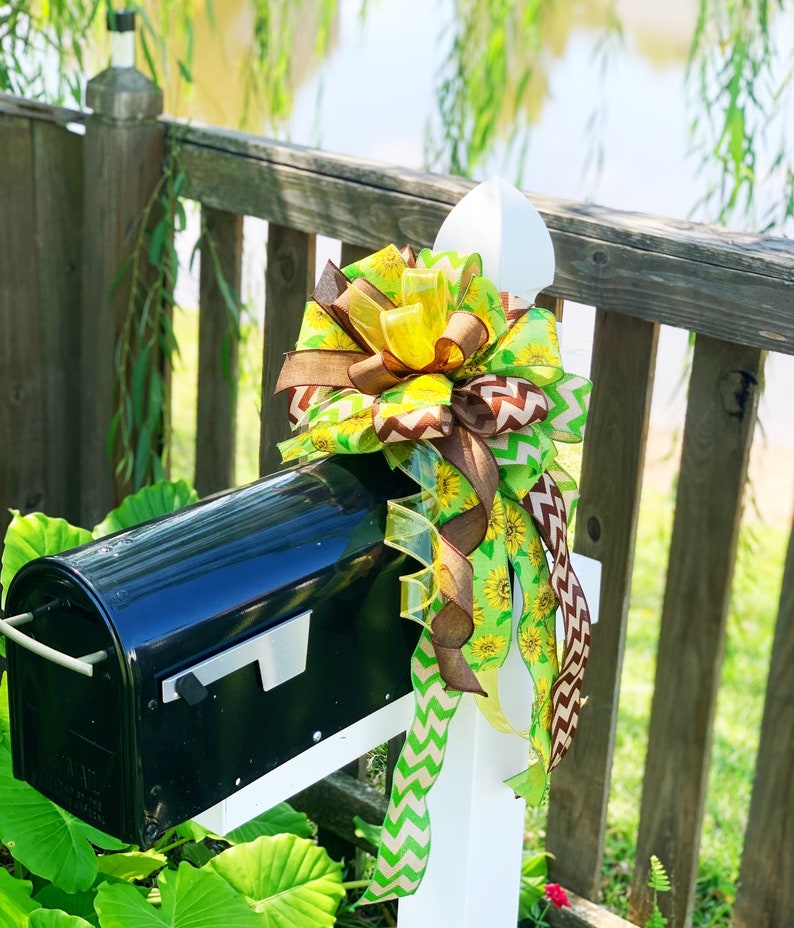 May include: A black mailbox with a decorative bow. The bow features yellow sunflowers, brown ribbons, and green chevron and solid ribbons. The mailbox is mounted on a white post, with a wooden fence and greenery in the background.