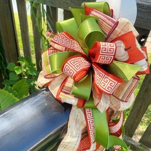 May include: A decorative bow with green, orange, and cream ribbons. The bow includes a pumpkin-themed ribbon and a ribbon with a geometric pattern. The bow is attached to a black surface, possibly a mailbox, in a garden setting.