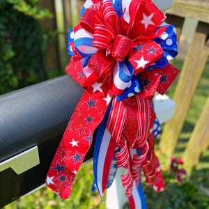 May include: A patriotic red, white, and blue bow with star and stripe patterns. The bow is made of multiple ribbons and is attached to a black surface. The bow is a decorative item, perfect for Independence Day or other patriotic holidays.
