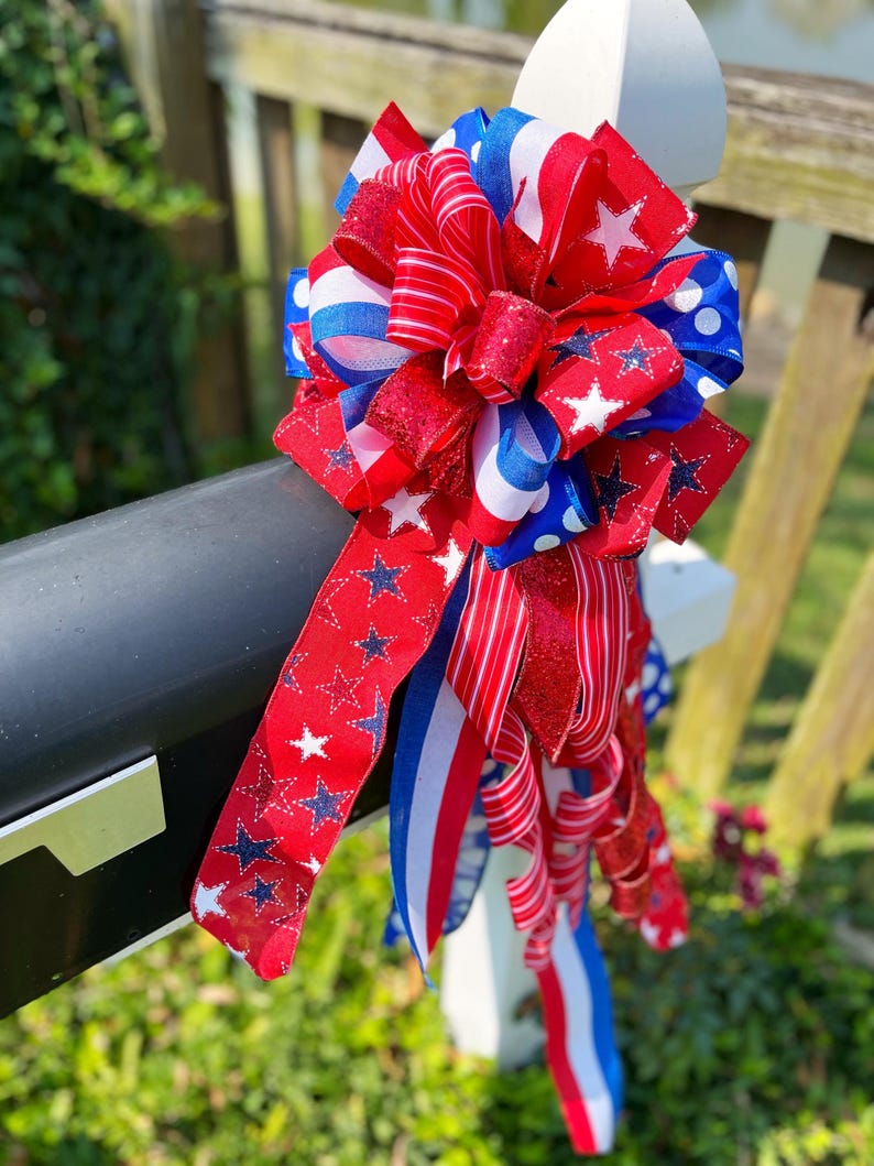 May include: A patriotic red, white, and blue bow with star and stripe patterns. The bow is made of multiple ribbons and is attached to a black surface. The bow is a decorative item, perfect for Independence Day or other patriotic holidays.