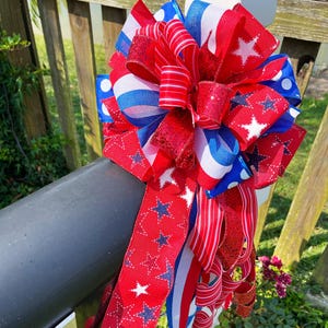 May include: A patriotic bow in red, white, and blue, featuring star and stripe patterns. The bow is made of various ribbons, including glittery and striped textures. It is attached to a black mailbox.