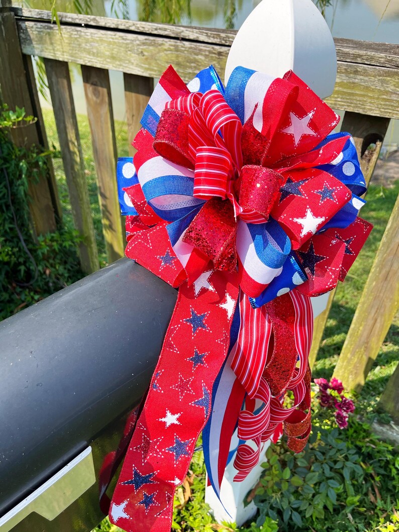 May include: A patriotic bow in red, white, and blue, featuring star and stripe patterns. The bow is made of various ribbons, including glittery and striped textures. It is attached to a black mailbox.