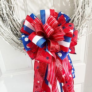 May include: A white twig wreath adorned with a large bow. The bow features red, white, and blue ribbons with star and polka dot patterns. Some ribbons have a glittery finish, adding a festive touch to the wreath.