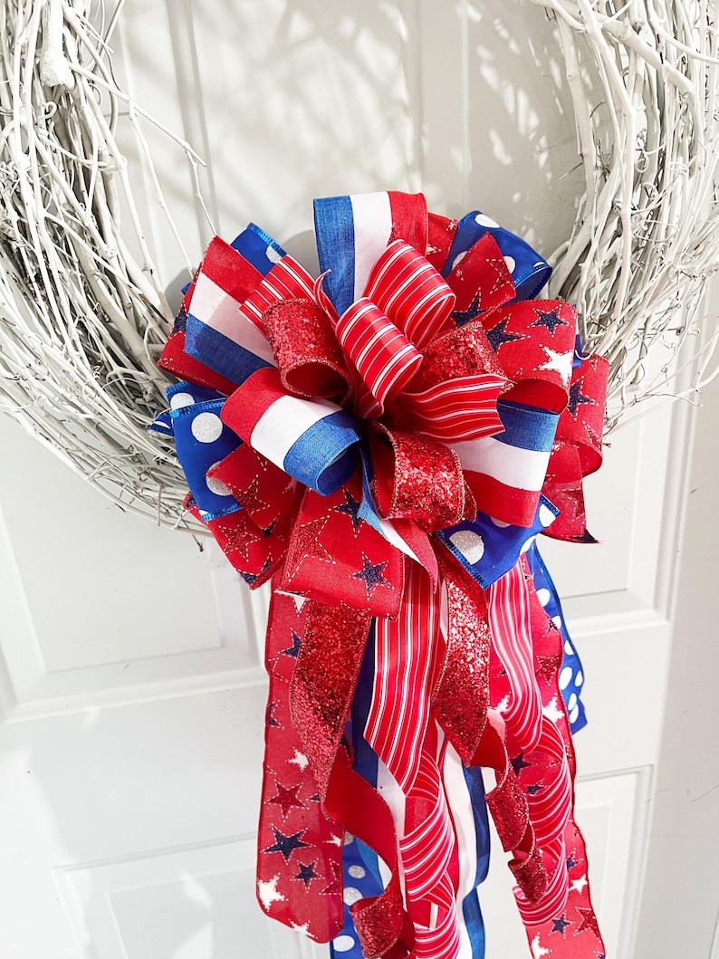 May include: A white twig wreath adorned with a large bow. The bow features red, white, and blue ribbons with star and polka dot patterns. Some ribbons have a glittery finish, adding a festive touch to the wreath.