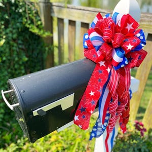 May include: A black mailbox adorned with a patriotic bow. The bow features red, white, and blue ribbons with star and stripe patterns. The mailbox is mounted on a white post, set against a backdrop of greenery and a wooden fence.