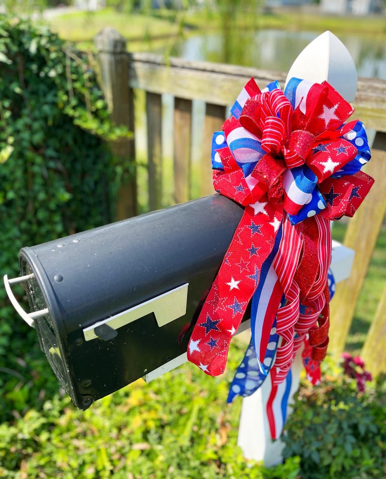 May include: A black mailbox adorned with a patriotic bow. The bow features red, white, and blue ribbons with star and stripe patterns. The mailbox is mounted on a white post, set against a backdrop of greenery and a wooden fence.