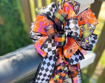 May include: A decorative Halloween bow featuring orange, black, and white ribbons. The bow includes ribbons with spiderwebs, checkerboard patterns, and the word "Halloween". The bow is tied to a black surface.