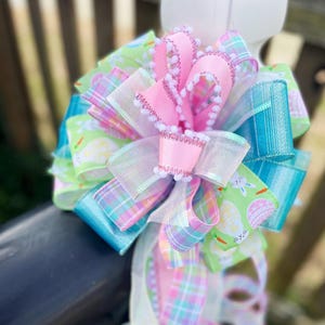 May include: A pastel pink, green, and blue Easter bow with a plaid pattern and a white pom-pom trim. The bow is decorated with a green ribbon featuring a pattern of Easter eggs and bunnies.