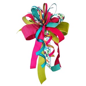 Puede incluir: Un lazo decorativo hecho de múltiples cintas en tonos vibrantes de rosa intenso, verde lima y turquesa, con una sección central que presenta un patrón floral multicolor sobre un fondo blanco, con largas cintas que cuelgan hacia abajo.
