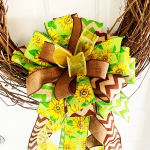 May include: A decorative wreath featuring a large bow with sunflower and chevron patterned ribbons in shades of yellow, green, and brown. The bow is attached to a natural grapevine wreath, creating a rustic and cheerful design.