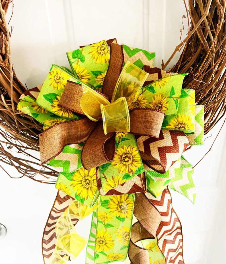 May include: A decorative wreath featuring a large bow with sunflower and chevron patterned ribbons in shades of yellow, green, and brown. The bow is attached to a natural grapevine wreath, creating a rustic and cheerful design.