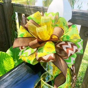May include: A decorative mailbox bow featuring a vibrant mix of ribbons. The bow incorporates sunflower and chevron patterns in shades of yellow, green, and brown. Clear, yellow ribbons add a touch of sparkle. The bow is attached to a black mailbox.