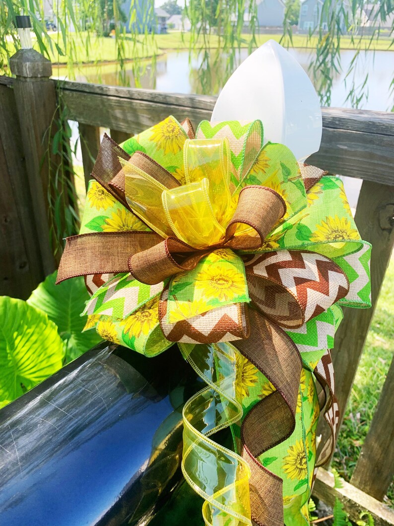 May include: A decorative mailbox bow featuring a vibrant mix of ribbons. The bow incorporates sunflower and chevron patterns in shades of yellow, green, and brown. Clear, yellow ribbons add a touch of sparkle. The bow is attached to a black mailbox.