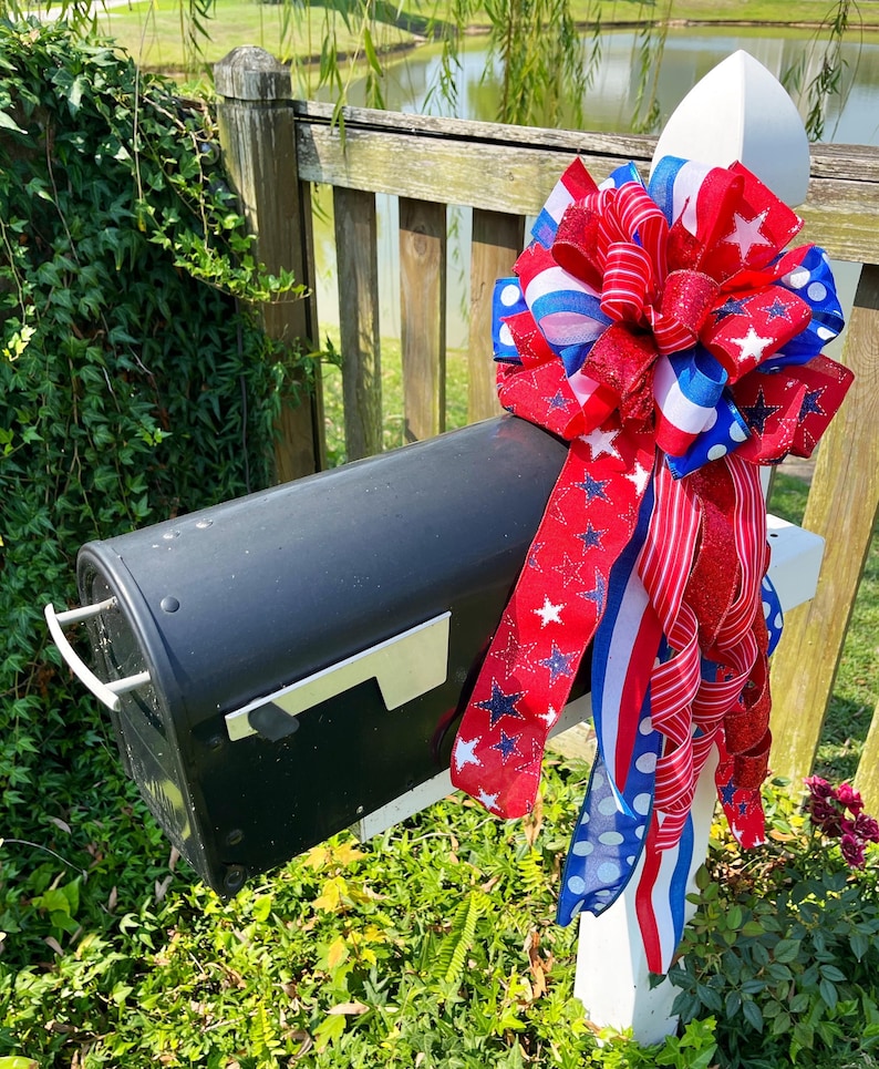 May include: A black mailbox with a decorative bow. The bow features red, white, and blue ribbons with star and stripe patterns. The mailbox is mounted on a white post, surrounded by greenery.
