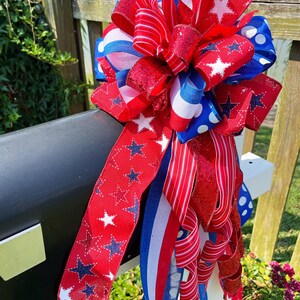 May include: A patriotic mailbox bow in red, white, and blue. The bow features various ribbon designs, including stars, stripes, and polka dots. The bow is attached to a black mailbox and white post, perfect for Independence Day.