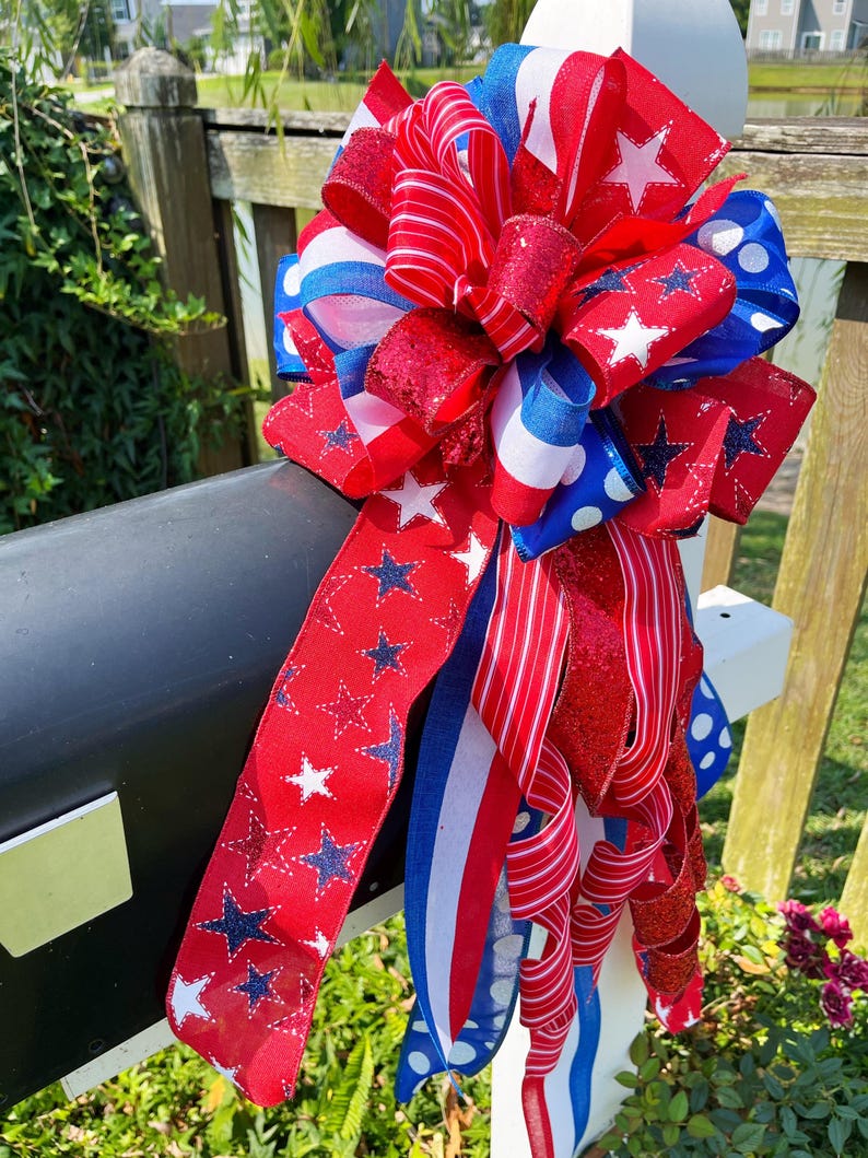 May include: A patriotic mailbox bow in red, white, and blue. The bow features various ribbon designs, including stars, stripes, and polka dots. The bow is attached to a black mailbox and white post, perfect for Independence Day.