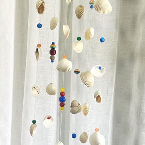 May include: A coastal-themed mobile featuring seashells, colorful beads, and decorative glass pieces. The mobile is suspended from a piece of driftwood and hangs in front of a white curtain. The beads are red, blue, yellow, and green.