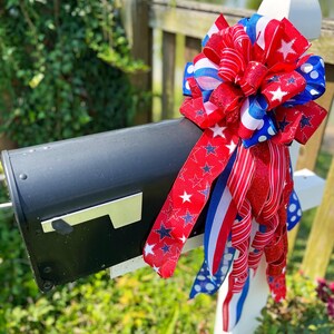 May include: A black mailbox adorned with a patriotic bow. The bow features red, white, and blue ribbons with star and stripe patterns. The mailbox is mounted on a white post, set against a backdrop of greenery and a blurred water feature.