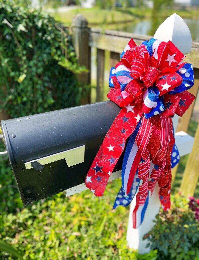 May include: A black mailbox adorned with a patriotic bow. The bow features red, white, and blue ribbons with star and stripe patterns. The mailbox is mounted on a white post, set against a backdrop of greenery and a blurred water feature.