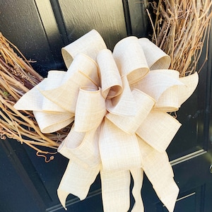 May include: A large, beige burlap bow tied on a grapevine wreath. The bow is tied with a large, loose knot and has long, flowing tails.