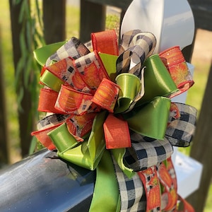 Könnte beinhalten: Eine große Schleife aus schwarz-weißem kariertem Band, grünem Satinband und orangefarbenem Band mit Kürbisdruck. Die Schleife ist um einen weißen Pfosten gebunden.