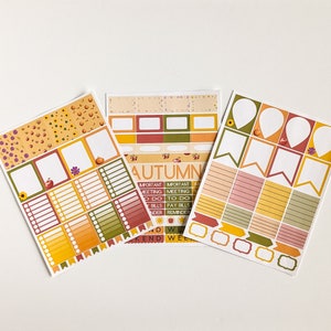 May include: Three sheets of planner stickers with an autumn theme. The stickers feature a variety of designs, including boxes, banners, and decorative elements in orange, yellow, green, and red. The word "AUTUMN" is printed on one sheet.