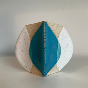 Op de afbeelding: Een geometrische keramische vaas met een witte, beige en blauwe glazuur. De vaas heeft een unieke vorm met een diamantvormig patroon.