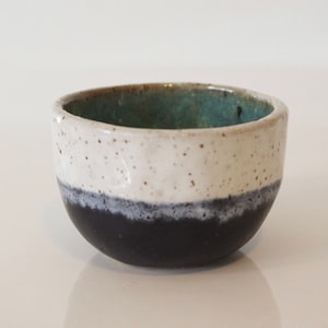 Pode incluir: Uma tigela pequena de cerâmica com um esmalte branco salpicado e uma base preta. O interior da tigela é esmaltado em uma cor azul esverdeada clara.