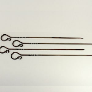May include: Set of four handcrafted metal skewers. Each skewer has a pointed end and a decorative looped handle. The metal has a dark, rustic finish. The skewers are arranged on a white background.
