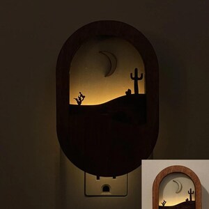 May include: A wooden night light with a cutout design of a desert scene with a crescent moon, cactus, and a small bush. The night light is plugged into a standard electrical outlet and is illuminated with a warm white light.