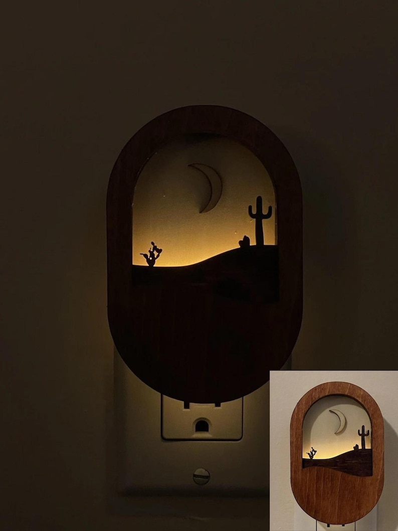 May include: A wooden night light with a cutout design of a desert scene with a crescent moon, cactus, and a small bush. The night light is plugged into a standard electrical outlet and is illuminated with a warm white light.