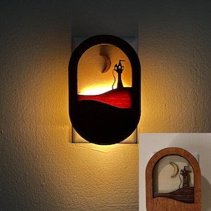 May include: A wooden night light with a silhouette of a cat in a tower with a crescent moon in the background. The night light is lit and casts a warm glow on the wall.