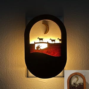 May include: A wooden night light with a crescent moon and goats silhouetted against a red and brown background. The night light is lit and glows warmly.