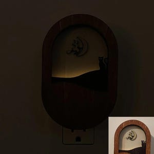 May include: A wooden night light with a silhouette of two cats and a crescent moon. The night light is plugged into an electrical outlet.