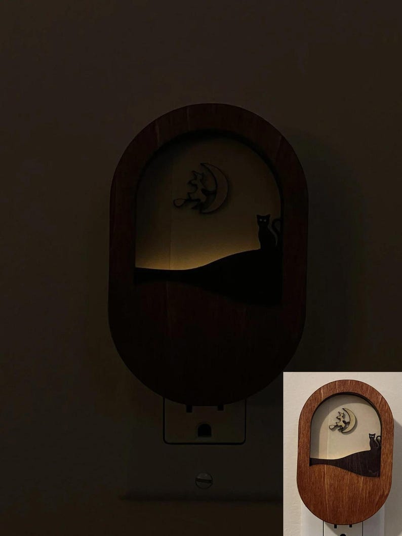 May include: A wooden night light with a silhouette of two cats and a crescent moon. The night light is plugged into an electrical outlet.