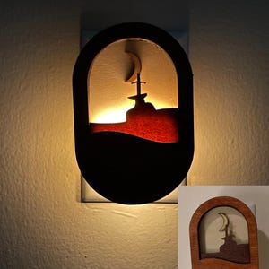 May include: A wooden night light with a crescent moon and a sword in a mountain silhouette. The night light is lit and glowing.