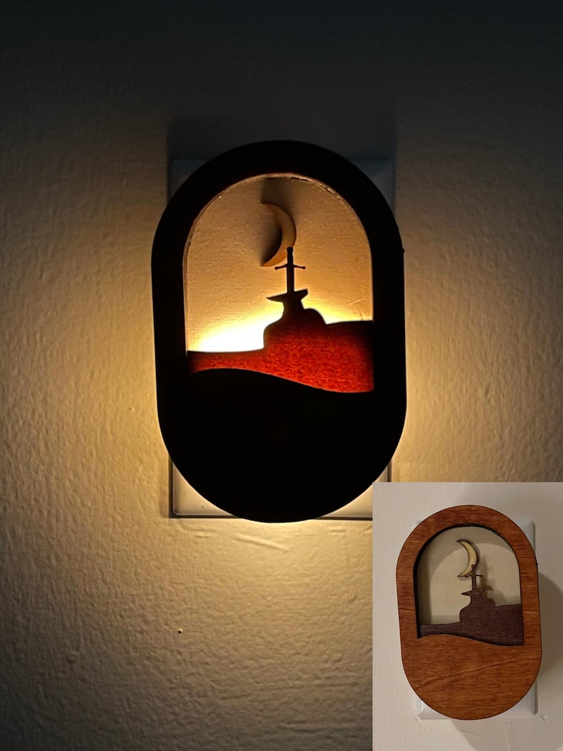 May include: A wooden night light with a crescent moon and a sword in a mountain silhouette. The night light is lit and glowing.