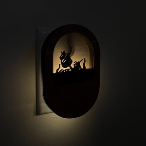 May include: A wooden night light shaped like an oval with a cutout design of a ship sailing on the ocean. The night light is plugged into an electrical outlet and emits a warm, yellow glow.