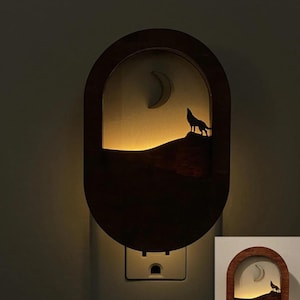 May include: A wooden night light with a silhouette of a wolf howling at the moon. The night light is plugged into an electrical outlet and is lit up.