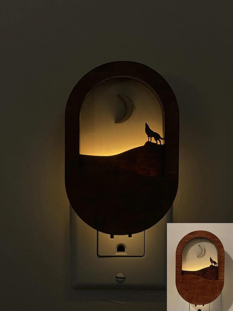 May include: A wooden night light with a silhouette of a wolf howling at the moon. The night light is plugged into an electrical outlet and is lit up.