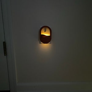 May include: A wooden night light plugged into an electrical outlet. The night light has a crescent moon and a mountain design. The light is on and illuminating the wall.