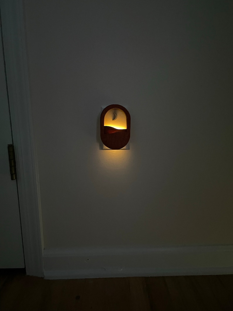 May include: A wooden night light plugged into an electrical outlet. The night light has a crescent moon and a mountain design. The light is on and illuminating the wall.