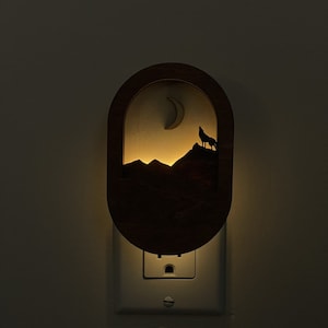 May include: A wooden night light featuring a silhouette of a wolf howling at the moon in front of a mountain range. The night light is plugged into an electrical socket.