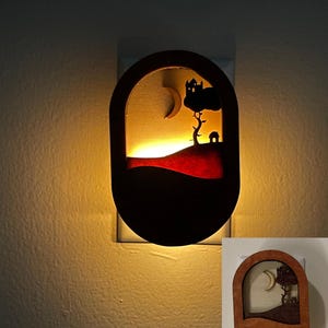 May include: A wooden night light shaped like an oval with a silhouette of a house, tree, and crescent moon. The night light is lit and glows a warm yellow.