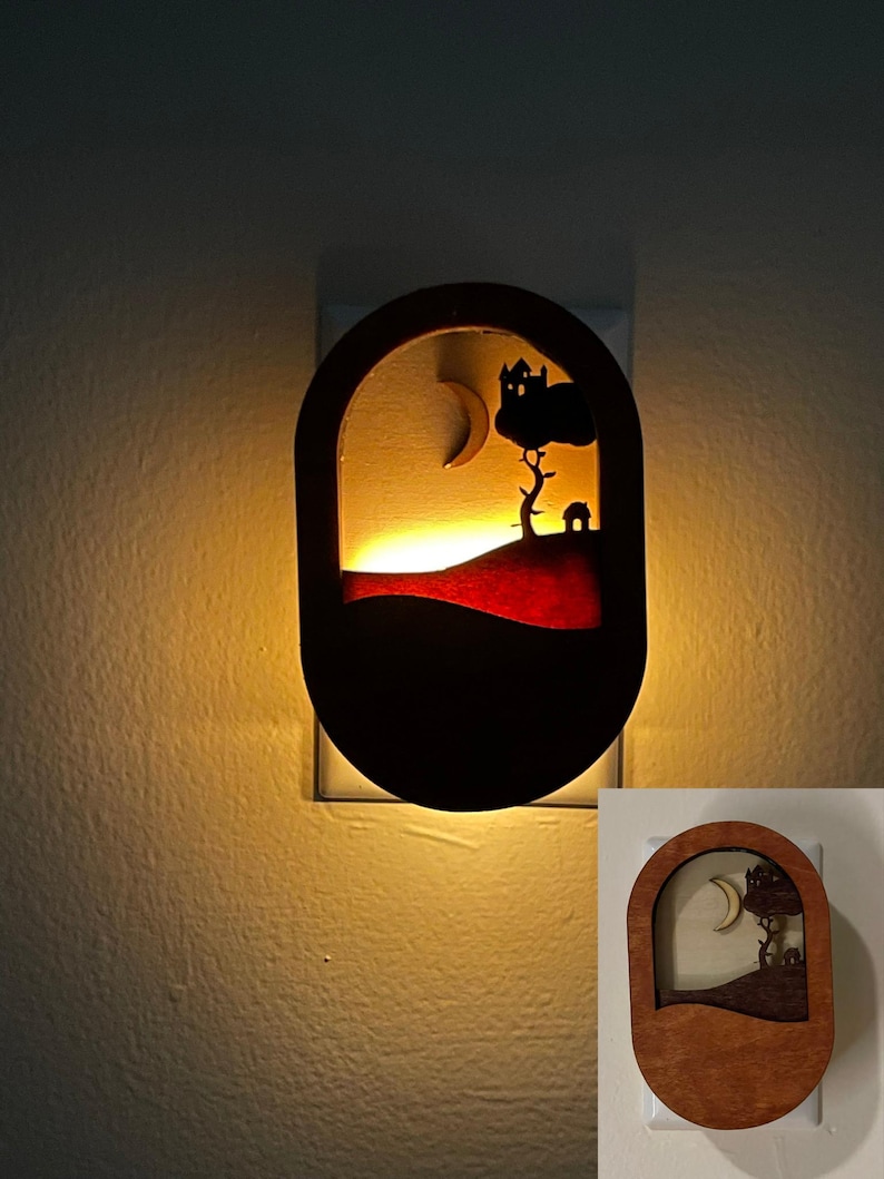 May include: A wooden night light shaped like an oval with a silhouette of a house, tree, and crescent moon. The night light is lit and glows a warm yellow.