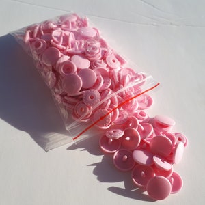 May include: A bag of pink plastic snap buttons. The buttons are in a clear plastic bag with a red zipper. The buttons are scattered on a white surface.
