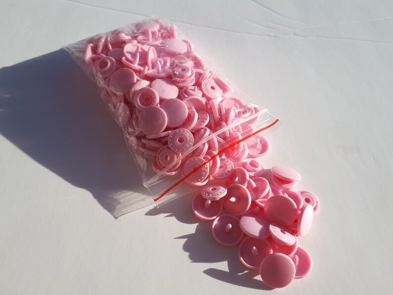 May include: A bag of pink plastic snap buttons. The buttons are in a clear plastic bag with a red zipper. The buttons are scattered on a white surface.